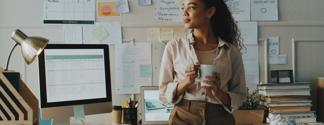 jeune fille adossée à son bureau au travail, un tasse à la main et qui regarde dehors en souriant