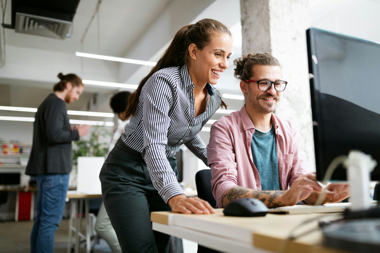 Deux collègues collaborent dans un bureau moderne : une femme debout sourit en regardant l’écran d’un ordinateur aux côtés d’un homme assis qui tape sur le clavier. D’autres personnes travaillent en arrière-plan dans un espace de travail lumineux et ouvert.