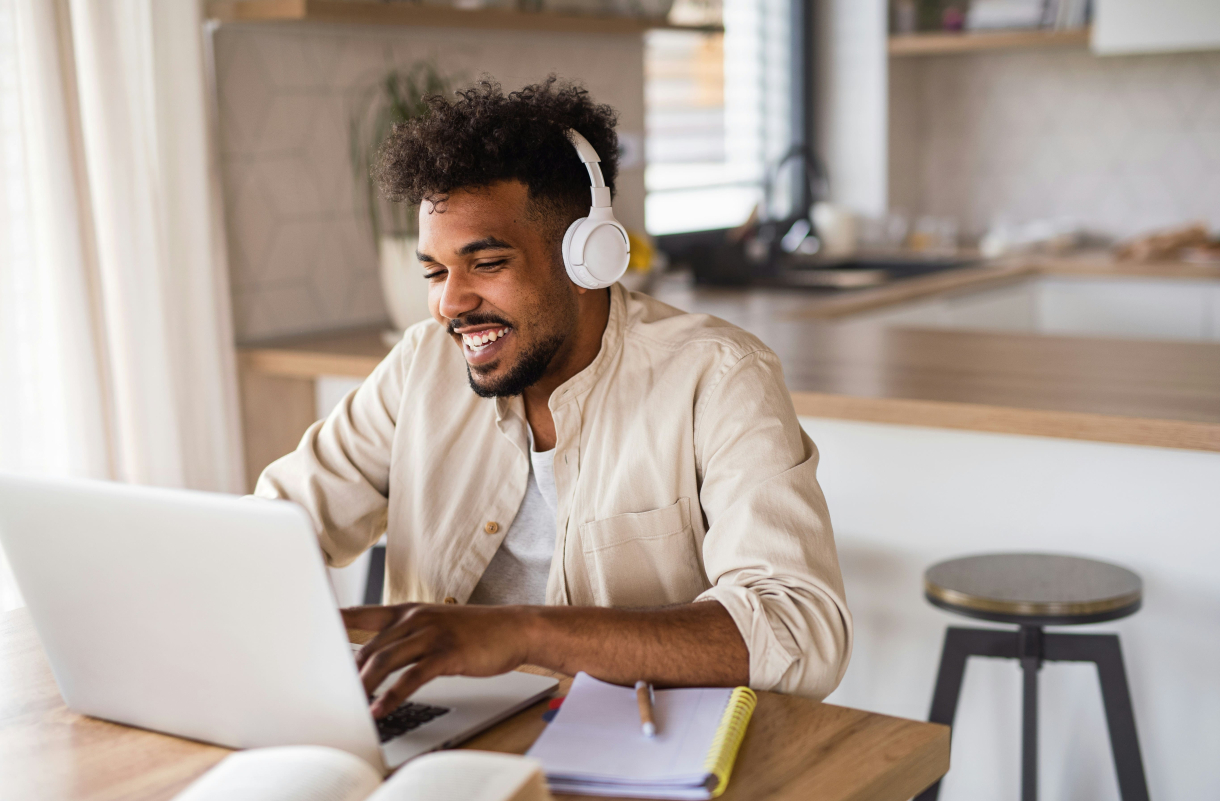 Un homme portant un casque audio blanc travaille sur un ordinateur portable dans un espace lumineux, avec un carnet et un stylo posés sur la table devant lui. Il sourit en regardant son écran.