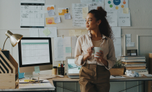 jeune fille adossée à son bureau au travail, un tasse à la main et qui regarde dehors en souriant